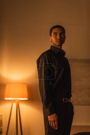 young african american man in black clothes looking away while standing in dark room with luminous lamp
