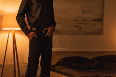 Téléchargez les photos : Vue partielle du mec afro-américain en vêtements noirs debout avec la main dans la poche dans la chambre la nuit - en image libre de droit