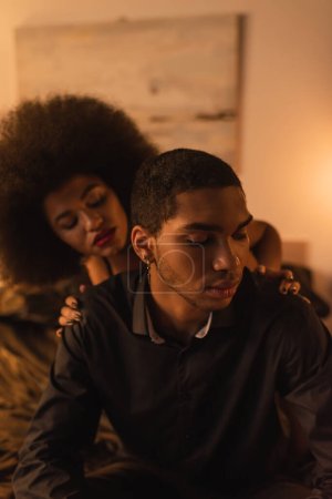 Foto de Mujer afroamericana rizada tocando hombros de hombre en camisa negra sentado en el dormitorio por la noche - Imagen libre de derechos