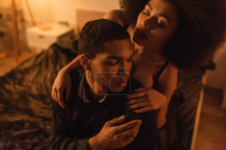 young african american woman with closed eyes embracing shoulders of boyfriend sitting on bed at home