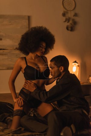 african american man hugging sexy young girlfriend in black underwear in dark bedroom with luminous lamp
