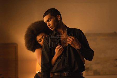 seductive african american woman undressing man in black shirt in dark bedroom with lighting