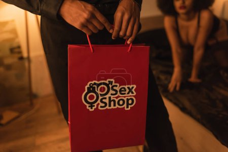 Photo for Cropped view of man holding paper bag from sex shop near african american girlfriend on blurred background - Royalty Free Image