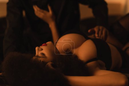 Foto de Passionate african american woman with sexy bust lying near blurred man on bed at home - Imagen libre de derechos