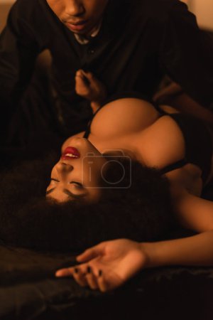 vue grand angle de la femme afro-américaine avec les yeux fermés et le sein sexy près du jeune homme sur le lit à la maison