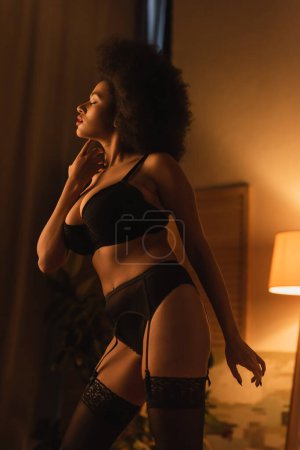 seductive african american woman in black underwear posing in dark bedroom with luminous lamp