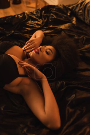 high angle view of brunette african american woman in black bra lying on silk bedding at home
