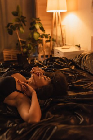 Téléchargez les photos : Jeune et sexy femme afro-américaine en soutien-gorge noir couché sur une literie en satin noir dans une chambre sombre avec éclairage - en image libre de droit