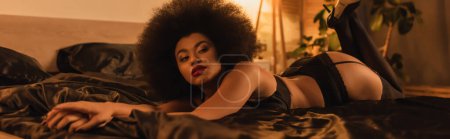 femme américaine africaine sensuelle en sous-vêtements sexy couché sur la literie de soie noire à la maison, bannière