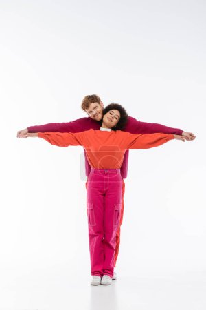 happy and interracial couple in magenta color clothes showing t letter on white background 