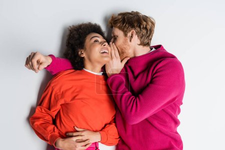 top view of redhead man in magenta color clothes whispering in ear of cheerful african american woman lying on white 