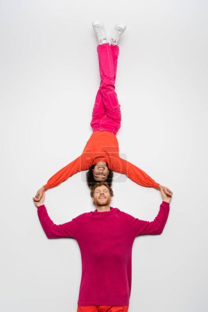Téléchargez les photos : Top view of happy redhead man and positive african american woman in magenta color clothes lying on white - en image libre de droit