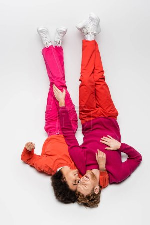top view of happy interracial couple in magenta color clothes lying upside down on white 