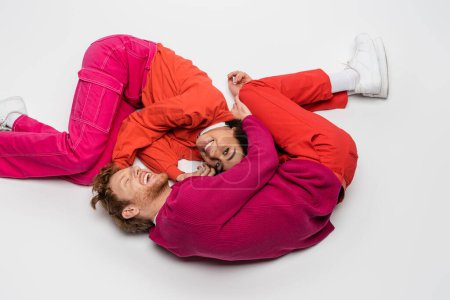 high angle view of smiling african american woman lying with redhead man in magenta color clothes on white