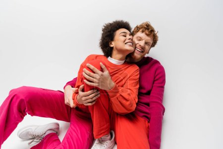 happy redhead man in magenta color sweatshirt hugging smiling african american woman on white 