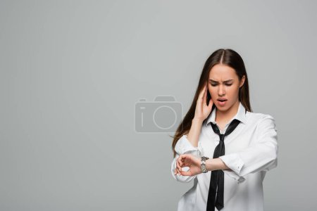 Téléchargez les photos : Femme choquée et brune en chemise blanche et cravate regardant montre bracelet isolé sur gris - en image libre de droit