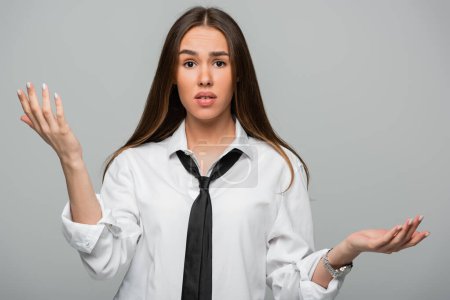 confused young woman in white shirt and tie gesturing isolated on grey