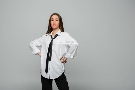 confident young woman in white shirt and tie looking at camera while standing with hands on hips isolated on grey 