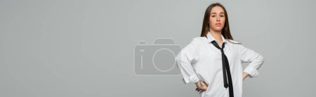 Foto de Confident young woman in white shirt and tie looking at camera while standing with hands on hips isolated on grey, banner - Imagen libre de derechos