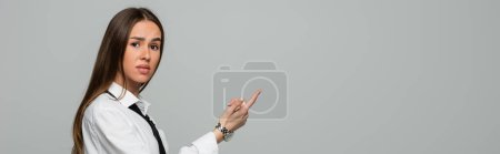 confused young woman in white shirt and tie pointing with finger isolated on grey, banner 