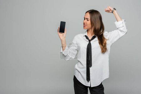 young woman in shirt and tie standing with clenched fist and holding smartphone with blank screen isolated on grey, gender equality