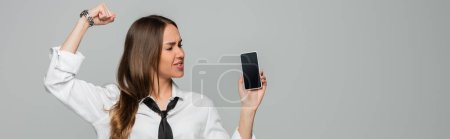 young woman in shirt and tie standing with clenched fist and holding smartphone with blank screen isolated on grey, banner 