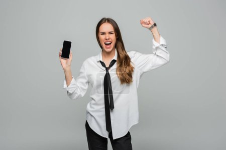 emotional woman in shirt and tie standing with clenched fist and holding smartphone with blank screen isolated on grey, gender equality