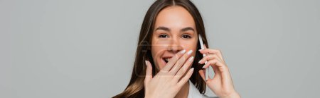 Téléchargez les photos : Femme heureuse parlant sur smartphone et souriant tout en couvrant la bouche isolée sur gris, bannière - en image libre de droit