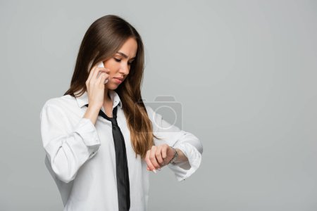 young woman in white shirt with tie talking on smartphone white looking at wristwatch isolated on grey 