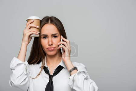 mujer joven en camisa blanca con corbata hablando en el teléfono inteligente y sosteniendo taza de papel con café para ir aislado en gris 
