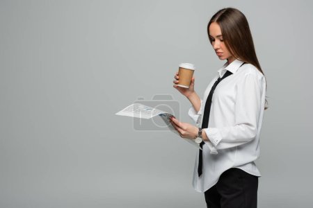 Photo for Young woman in white shirt with tie reading newspaper while holding paper cup isolated on grey - Royalty Free Image