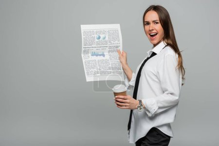 Photo for Amazed young woman in white shirt with tie holding newspaper and paper cup isolated on grey - Royalty Free Image