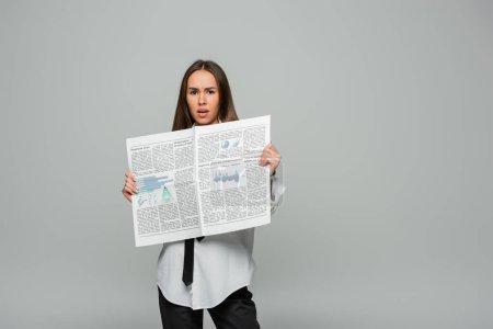 shocked young woman in white shirt with tie holding newspaper while looking at camera isolated on grey 