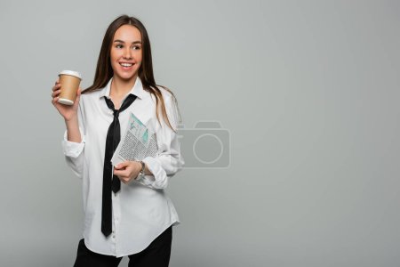 Photo for Cheerful woman in white shirt with tie holding newspaper and paper cup with coffee to go isolated on grey - Royalty Free Image