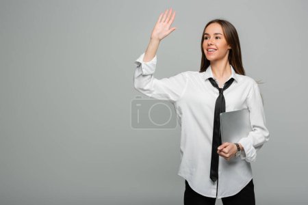 Téléchargez les photos : Femme heureuse en chemise blanche avec cravate tenant ordinateur portable tout en agitant la main isolée sur gris - en image libre de droit