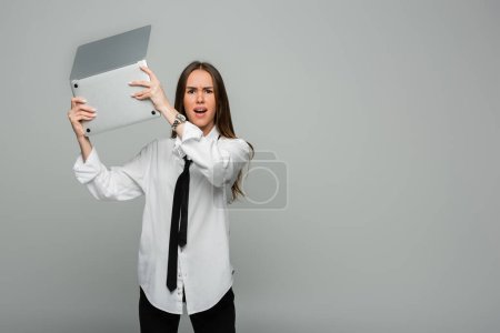 Wütende junge Frau im weißen Hemd mit Krawatte hält Laptop isoliert auf grau 