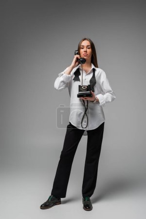 Foto de Full length of young woman in white shirt with tie and pants using retro telephone on grey - Imagen libre de derechos