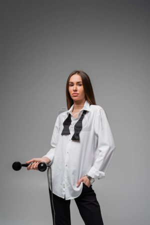 junge Frau in weißem Hemd mit Krawatte und Hose hält Retro-Telefon in der Hand, während sie auf grau posiert 