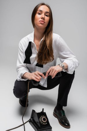 Foto de Brunette woman in white shirt with tie and pants holding retro telephone while sitting on grey - Imagen libre de derechos