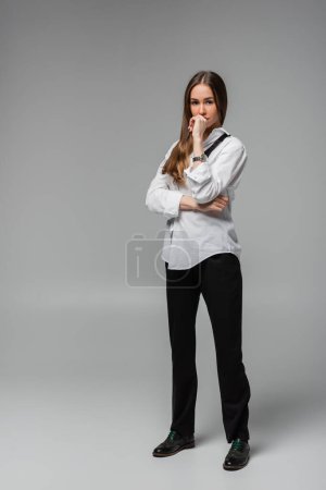 full length of pensive woman in shirt with tie and black pants standing on grey