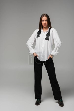 longitud completa de la mujer confiada en camisa blanca con corbata y pantalones de pie con las manos en los bolsillos en gris, concepto de igualdad de género 