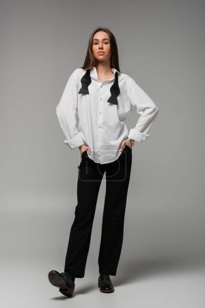 full length of confident woman in white shirt and trousers standing with hands in pockets on grey, gender equality concept 