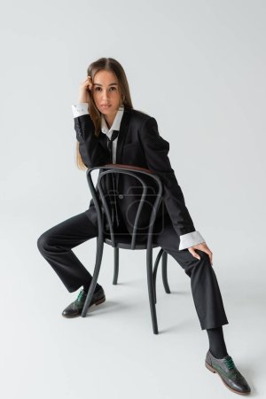 volle Länge der selbstbewussten Frau im Anzug sitzt auf einem Holzstuhl auf grau 