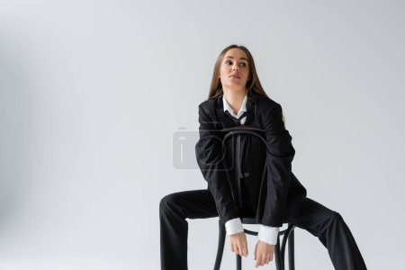young and brunette woman in suit sitting on wooden chair on grey background 