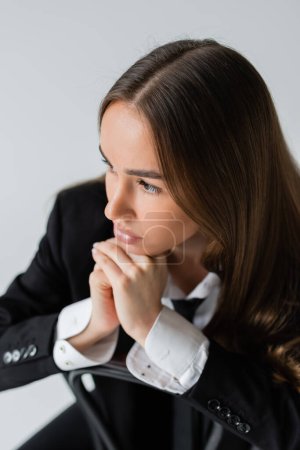 Photo for High angle view of young and brunette woman in suit leaning on wooden chair back isolated on grey - Royalty Free Image