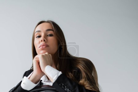 Foto de Low angle view of brunette woman with long hair sitting in suit and leaning on wooden chair back isolated on grey - Imagen libre de derechos