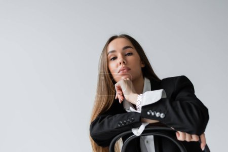 Photo for Confident woman with long hair sitting in suit and leaning on wooden chair back isolated on grey - Royalty Free Image