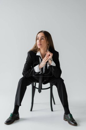 full length of young brunette woman in black suit with tie sitting on wooden chair and looking away on grey 
