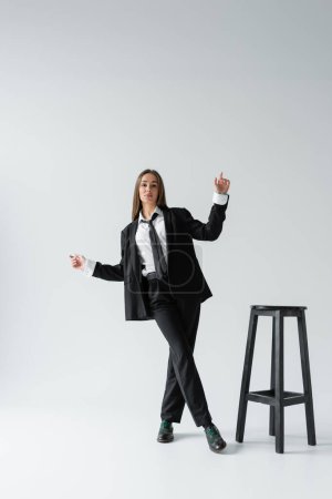 Téléchargez les photos : Pleine longueur de jeune femme brune en costume noir avec cravate posant près de chaise haute sur gris - en image libre de droit