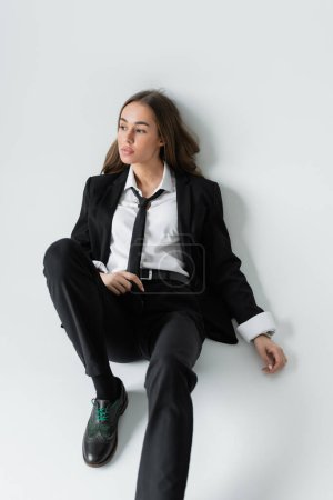 full length of pensive woman in black formal wear with tie sitting on grey background 
